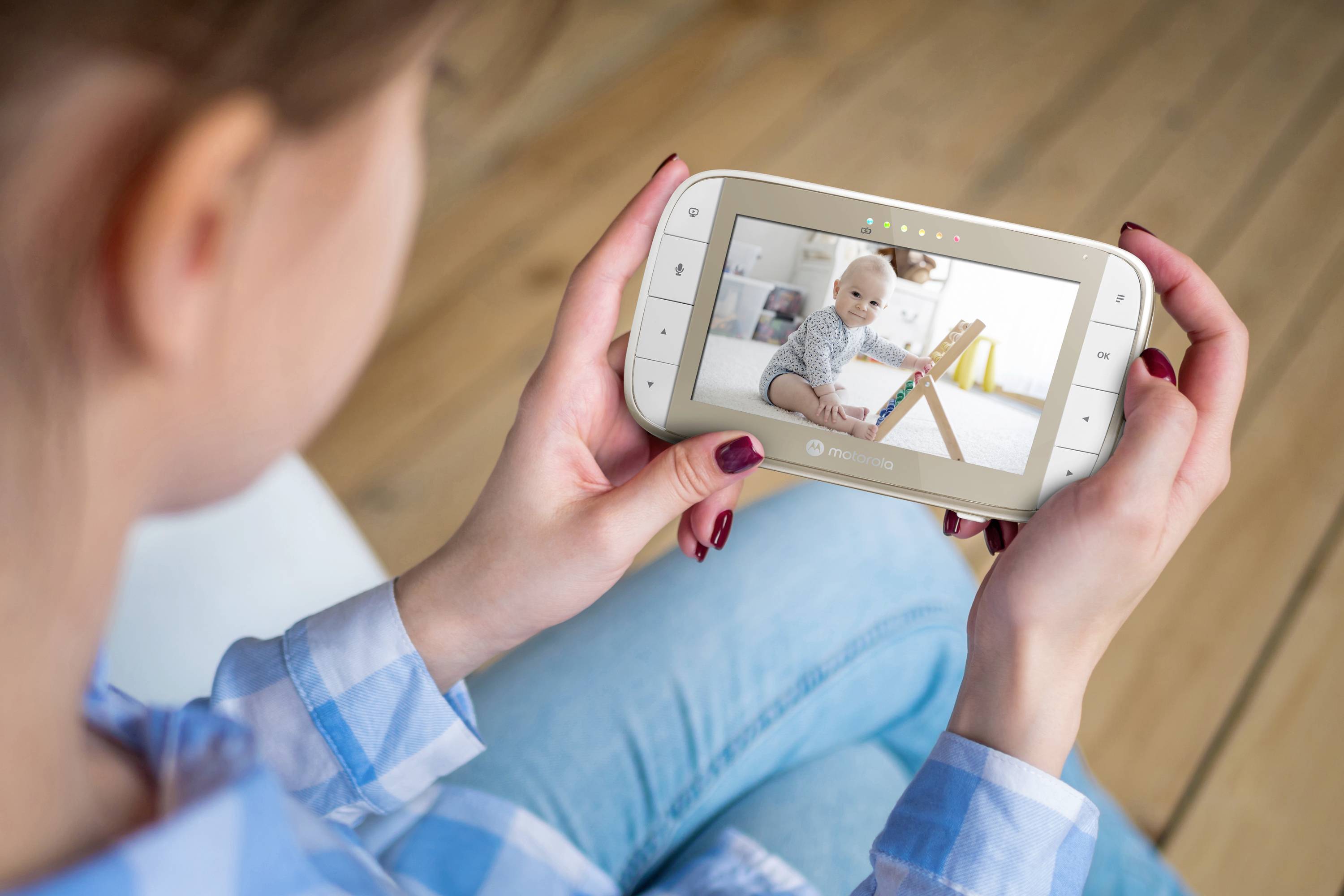 A woman holds a baby monitor showing a baby playing on the floor with a small wooden toy.