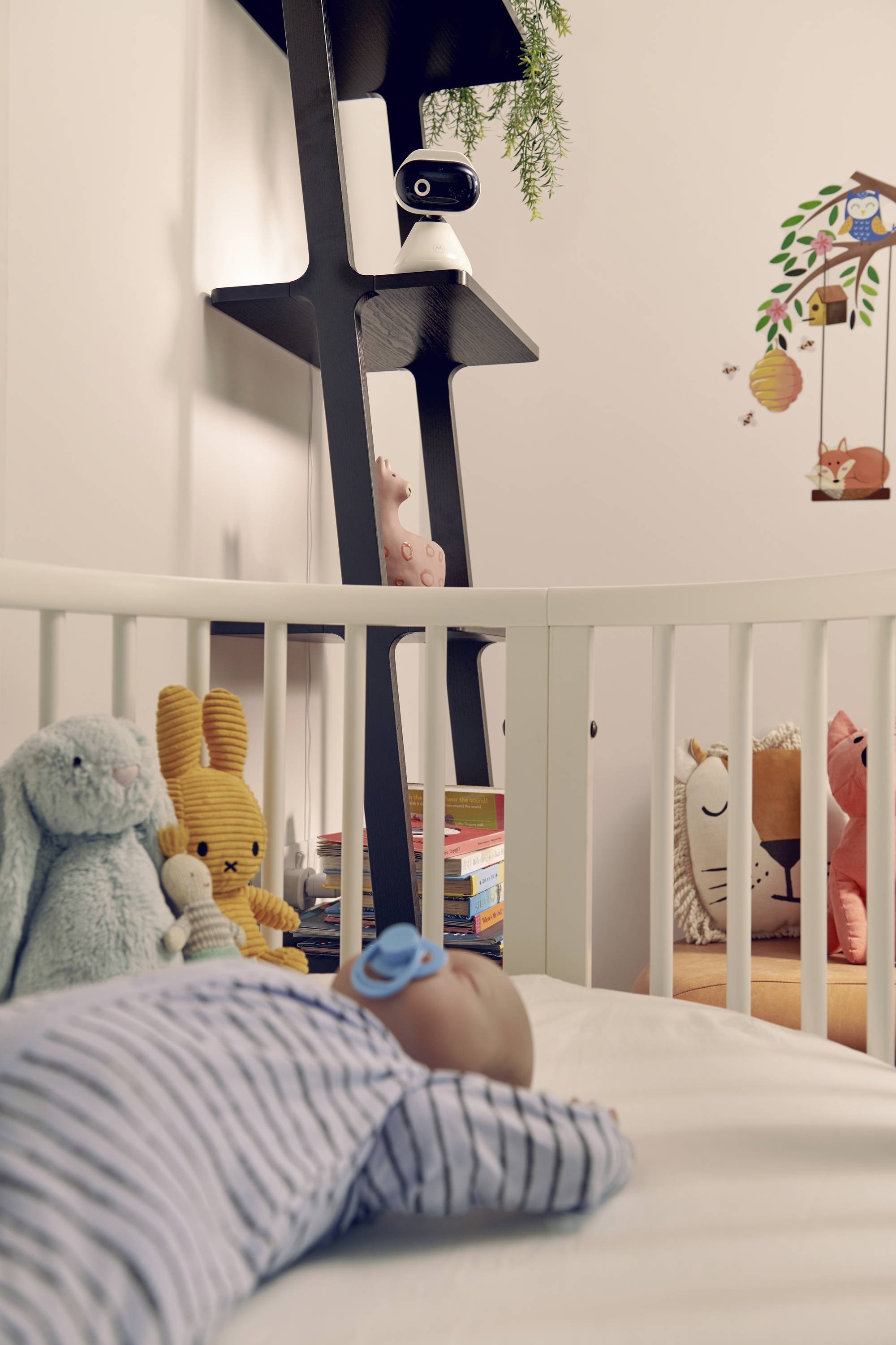 A baby sleeps in a crib, surrounded by plush toys. A black shelf with books and a camera overlooks the scene. Wall has animal decals.