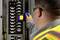A technician wearing a safety vest uses a thermal camera to inspect an electrical panel for temperature irregularities.