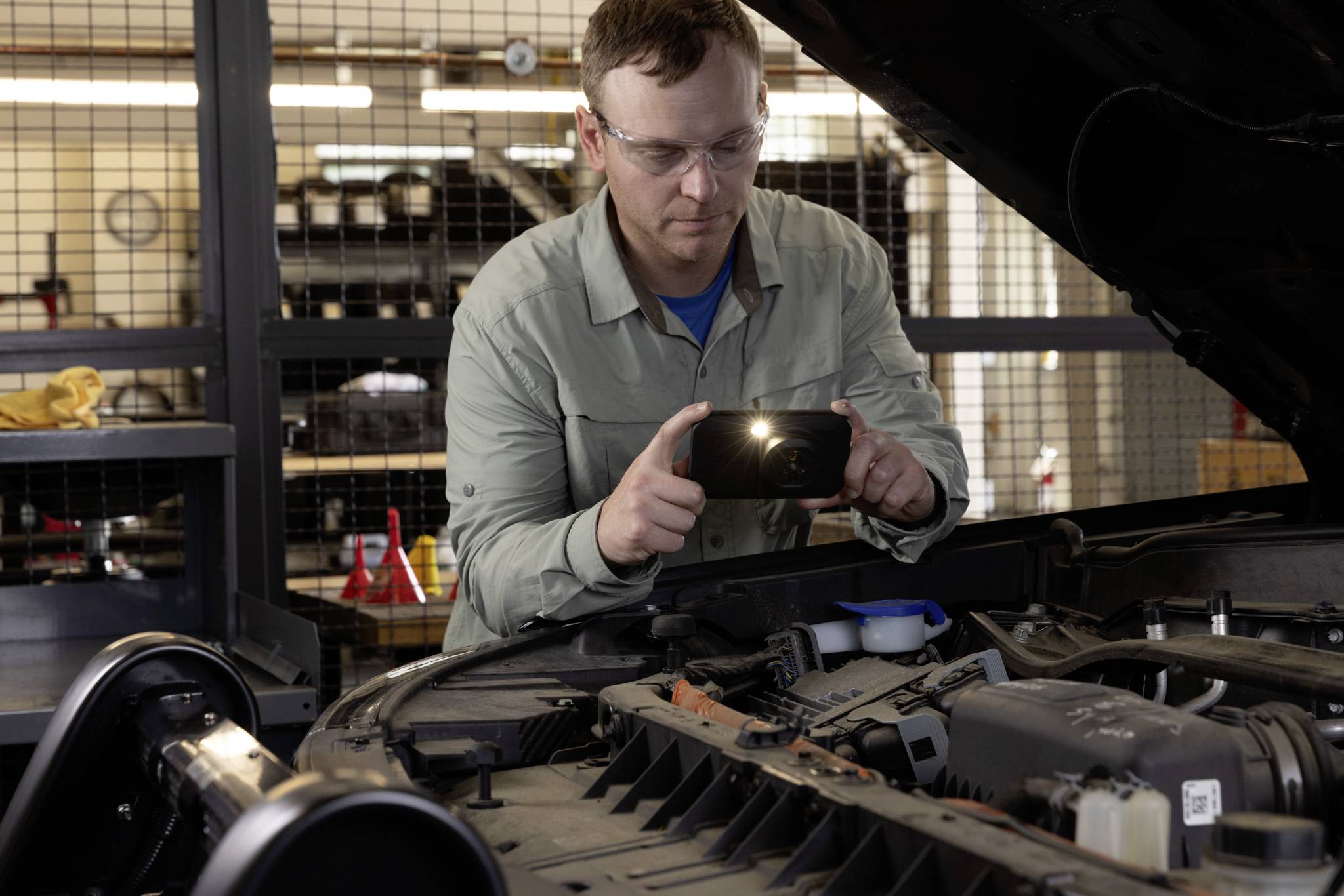 A mechanic wearing safety glasses inspects a car engine closely while using a flashlight, inside a well-lit workshop.