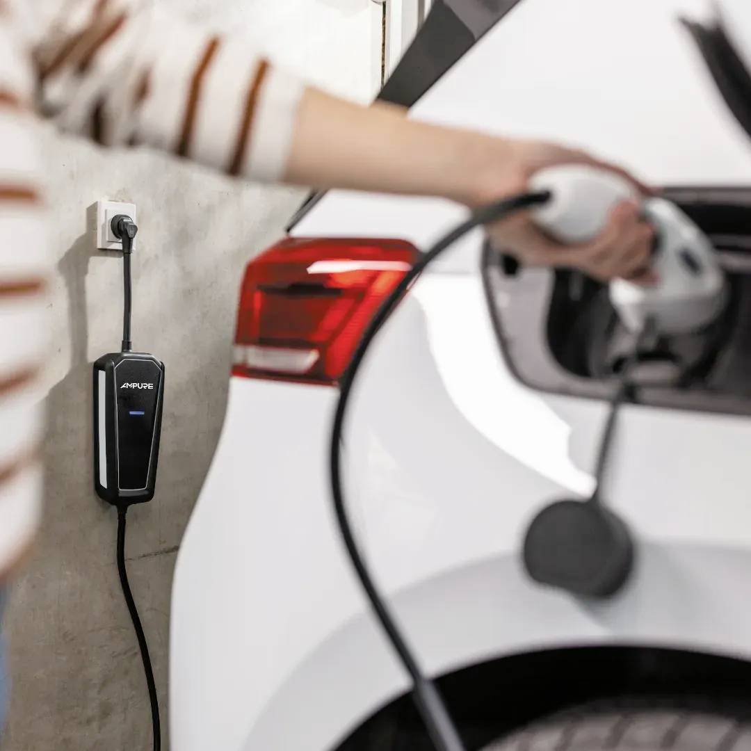A white electric vehicle is charging in a garage, plugged into a wall-mounted charger. An individual is holding the charger handle.
