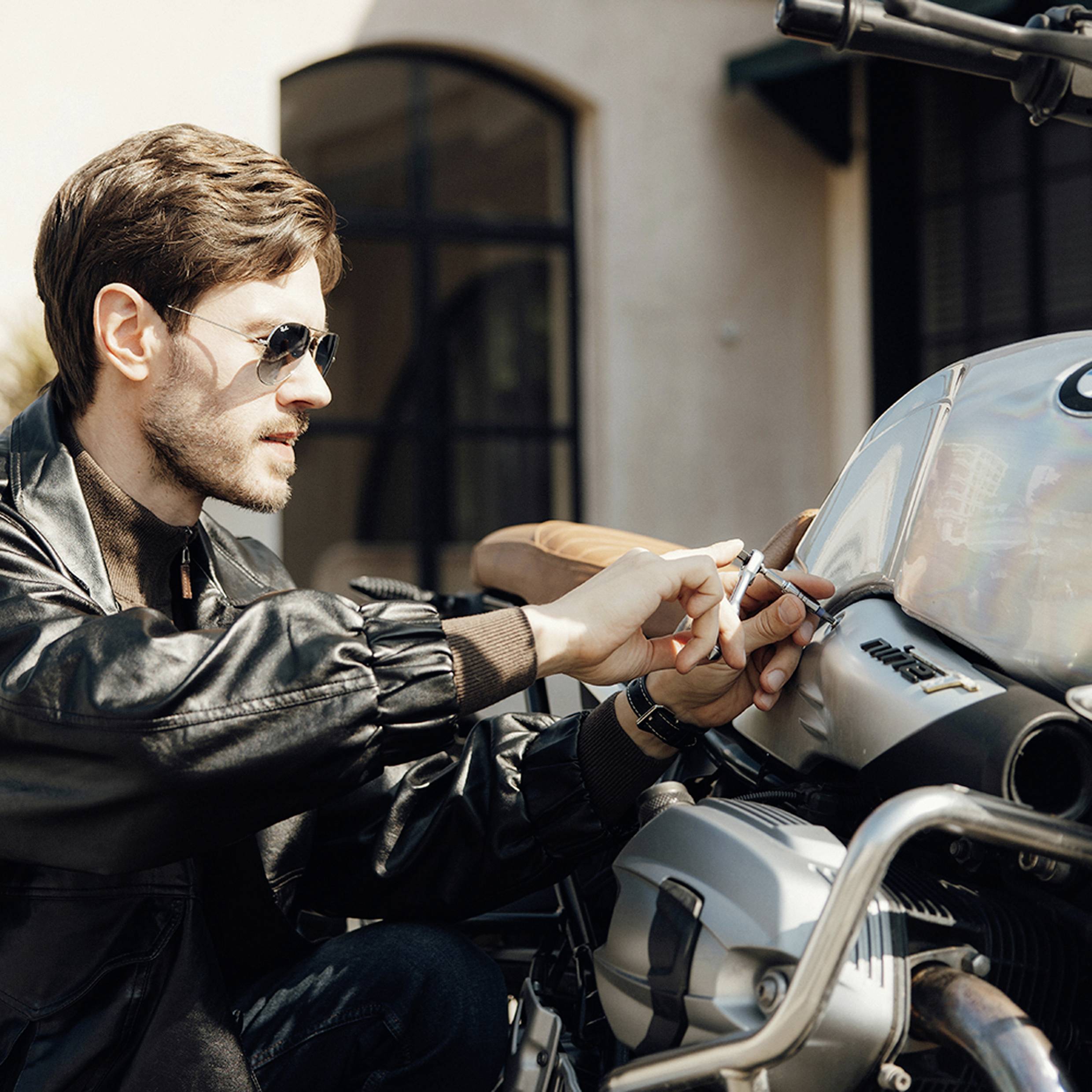 A man wearing sunglasses and a leather jacket is repairing or adjusting a motorcycle. The setting is outdoors in daylight near a building.