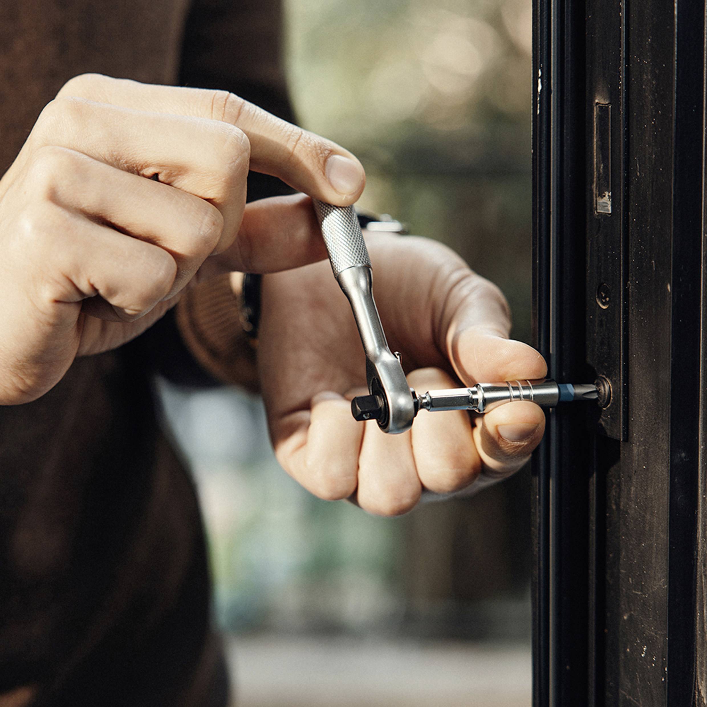 Person using a wrench to adjust or fix a door hinge, focusing on hands and tool. The background is blurred, indicating an indoor setting.