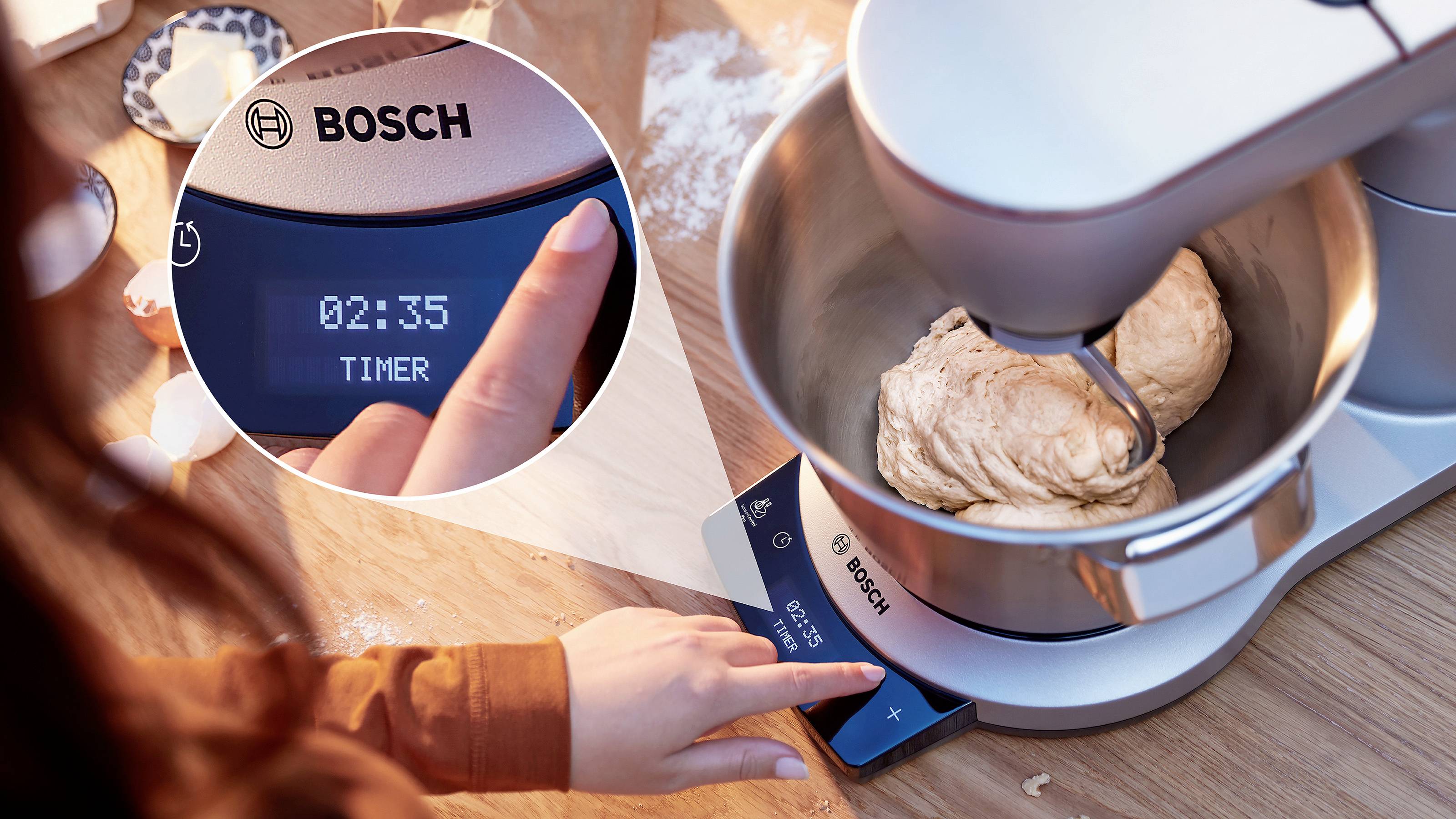 A person sets a Bosch stand mixer's timer to 2 minutes and 35 seconds while kneading dough. The timer is highlighted in a close-up view.