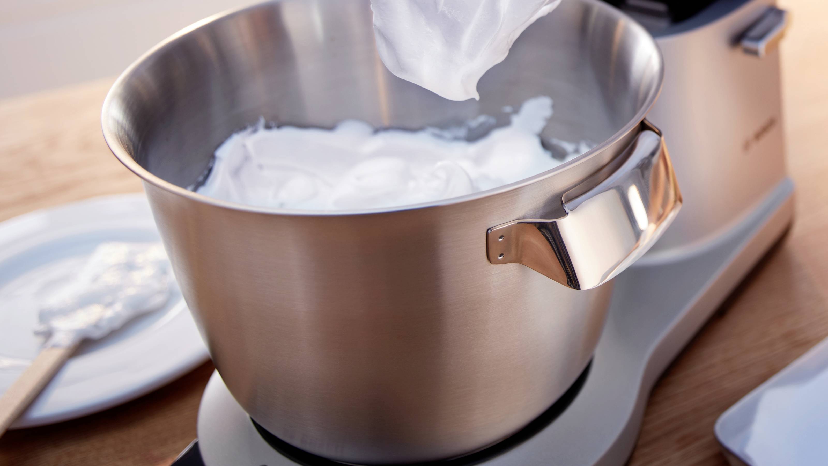 A bowl of whipped cream on a mixer, with a beater lifting some of the cream. A plate and spatula are nearby on a wooden surface.