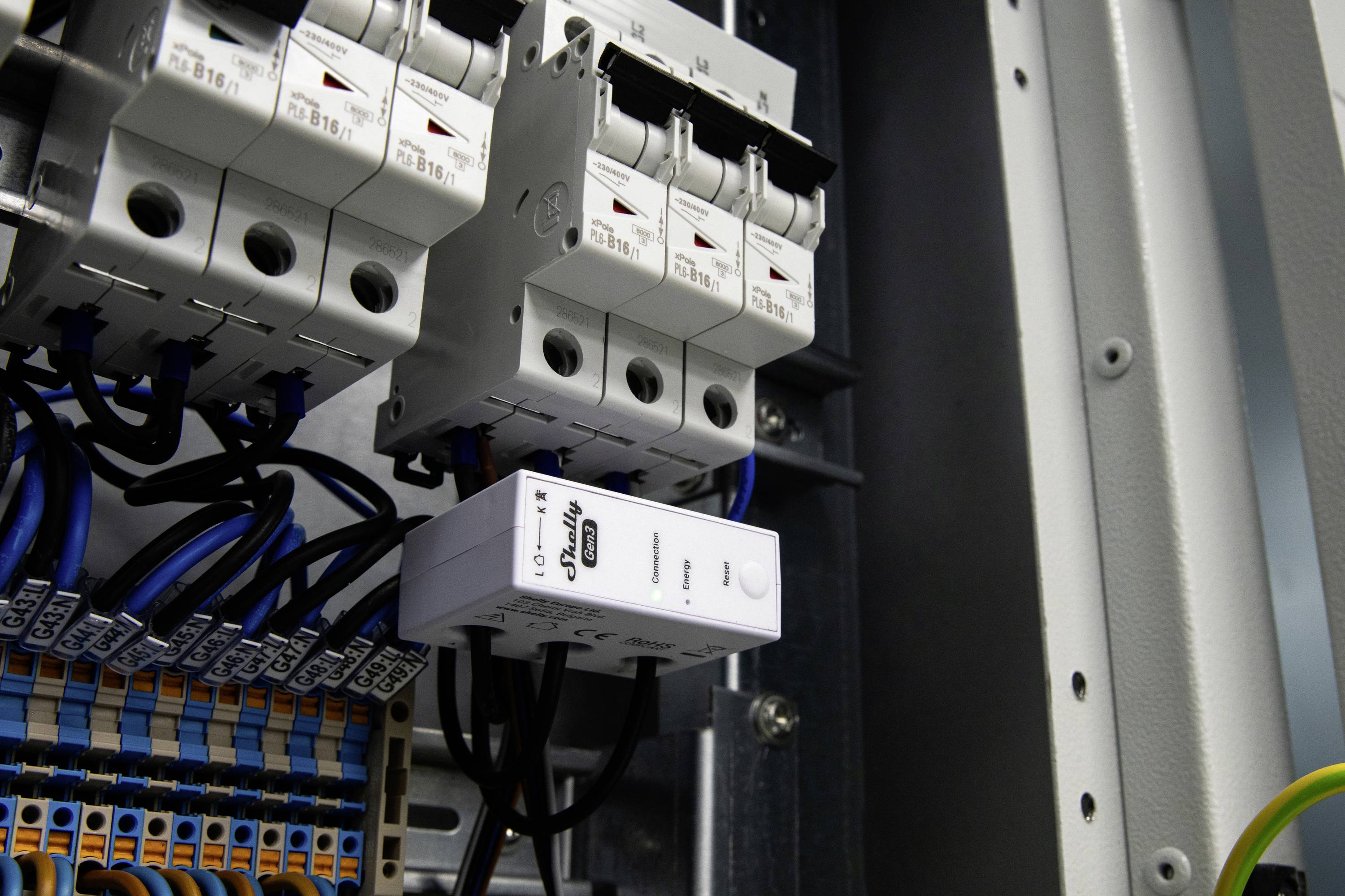 An electrical circuit breaker panel with multiple switches and wires, including a digital energy meter, is shown inside an industrial setting.