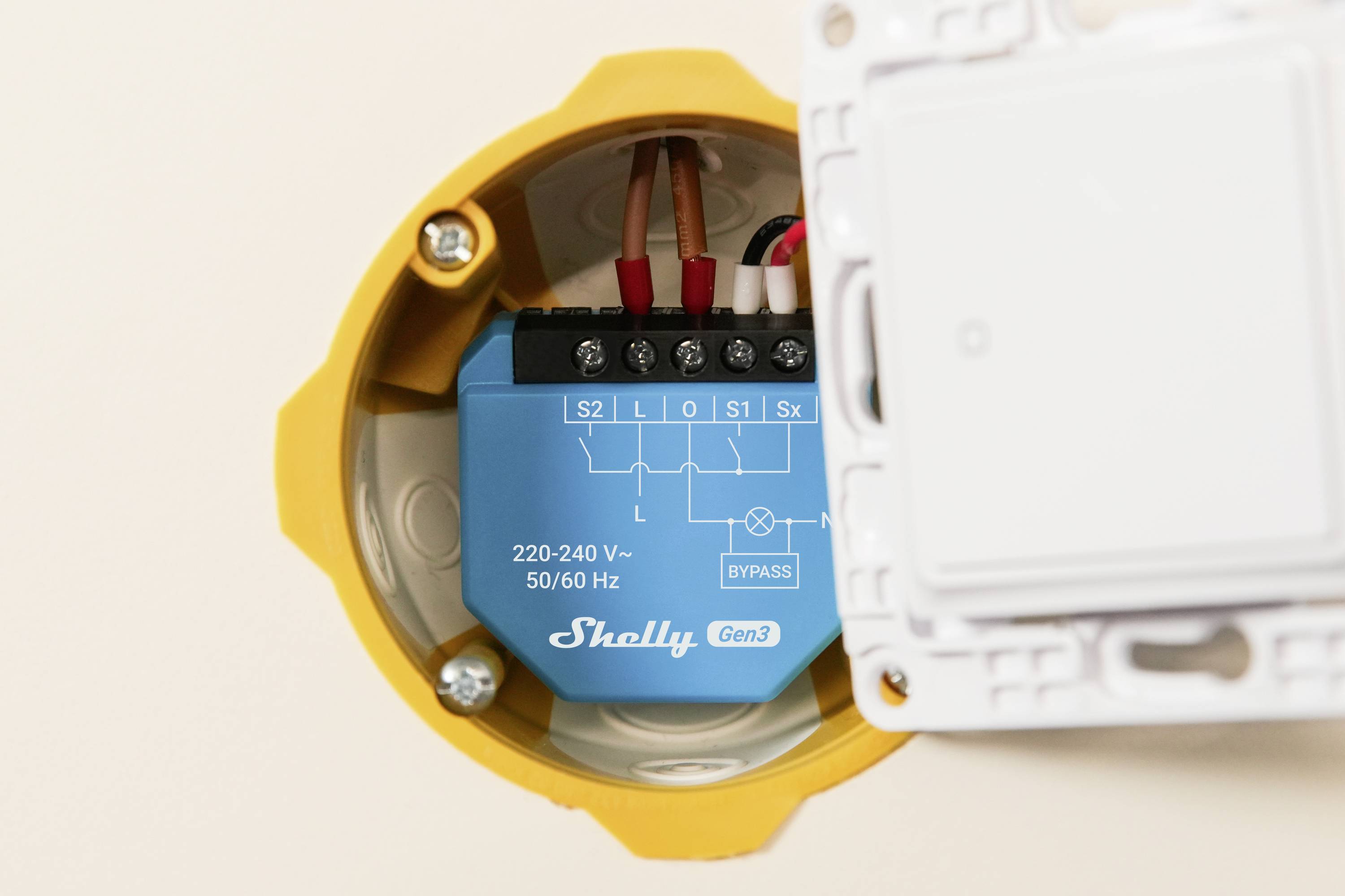 A blue Shelly Gen3 device installed inside a yellow electrical box, showing wiring connections labeled S1, S2, L, O, I, with voltage details.