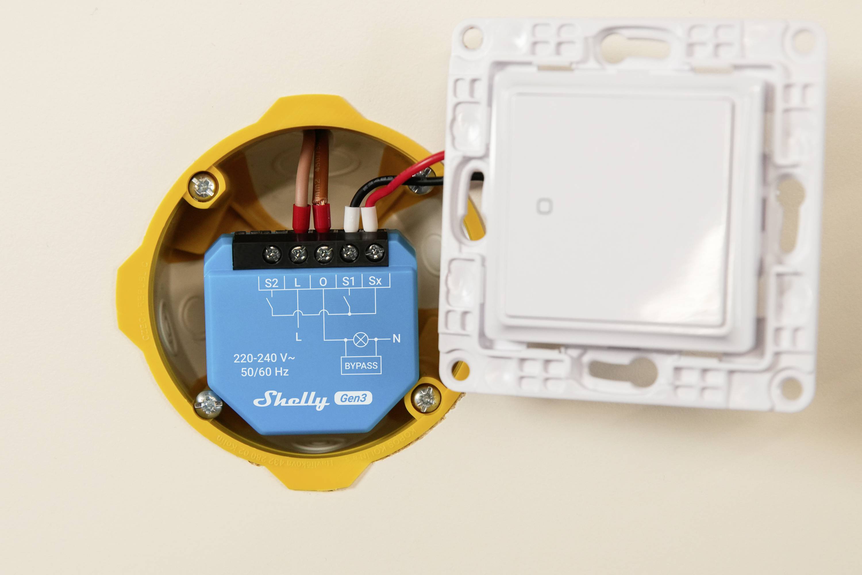 An open wall switch box showing internal wiring and a Shelly Gen 1 device for controlling power, labeled with voltage specifications.