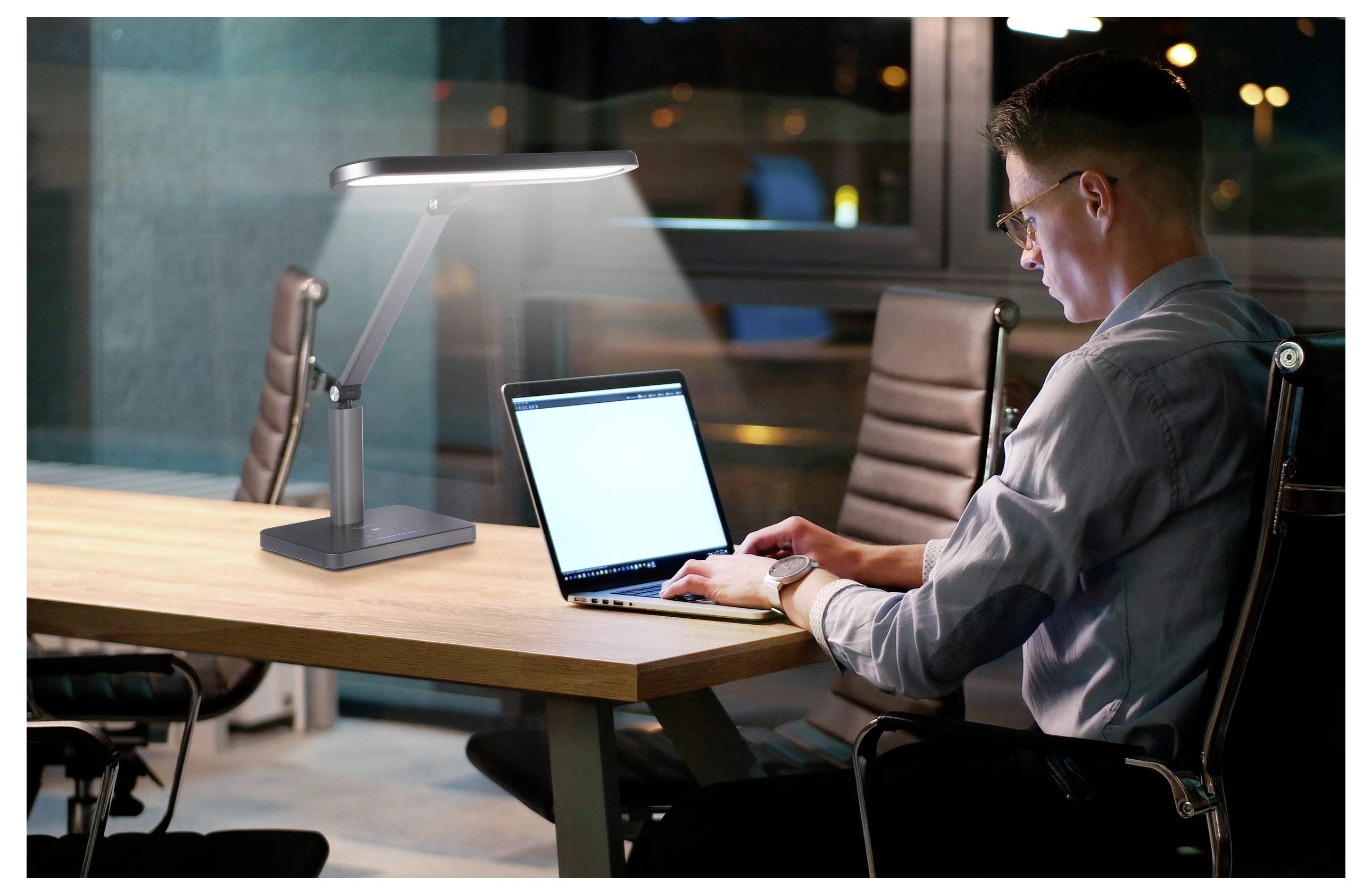 A man in an office works on a laptop, illuminated by a desk lamp, with large windows and city lights visible in the background.