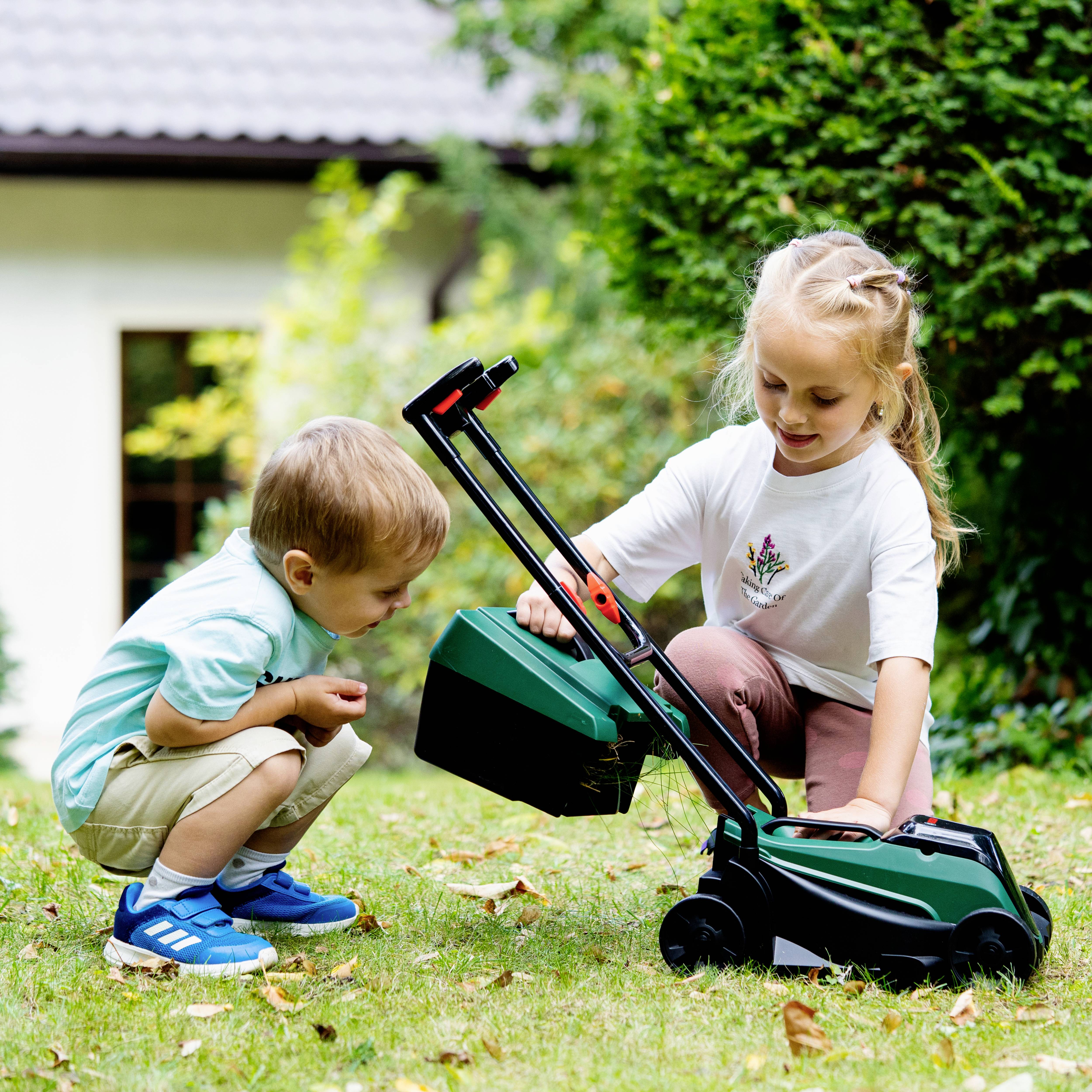 Two young children play outside with a toy lawnmower on grass; a house is partially visible in the background.