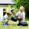 Two young children play outside with a toy lawnmower on grass; a house is partially visible in the background.