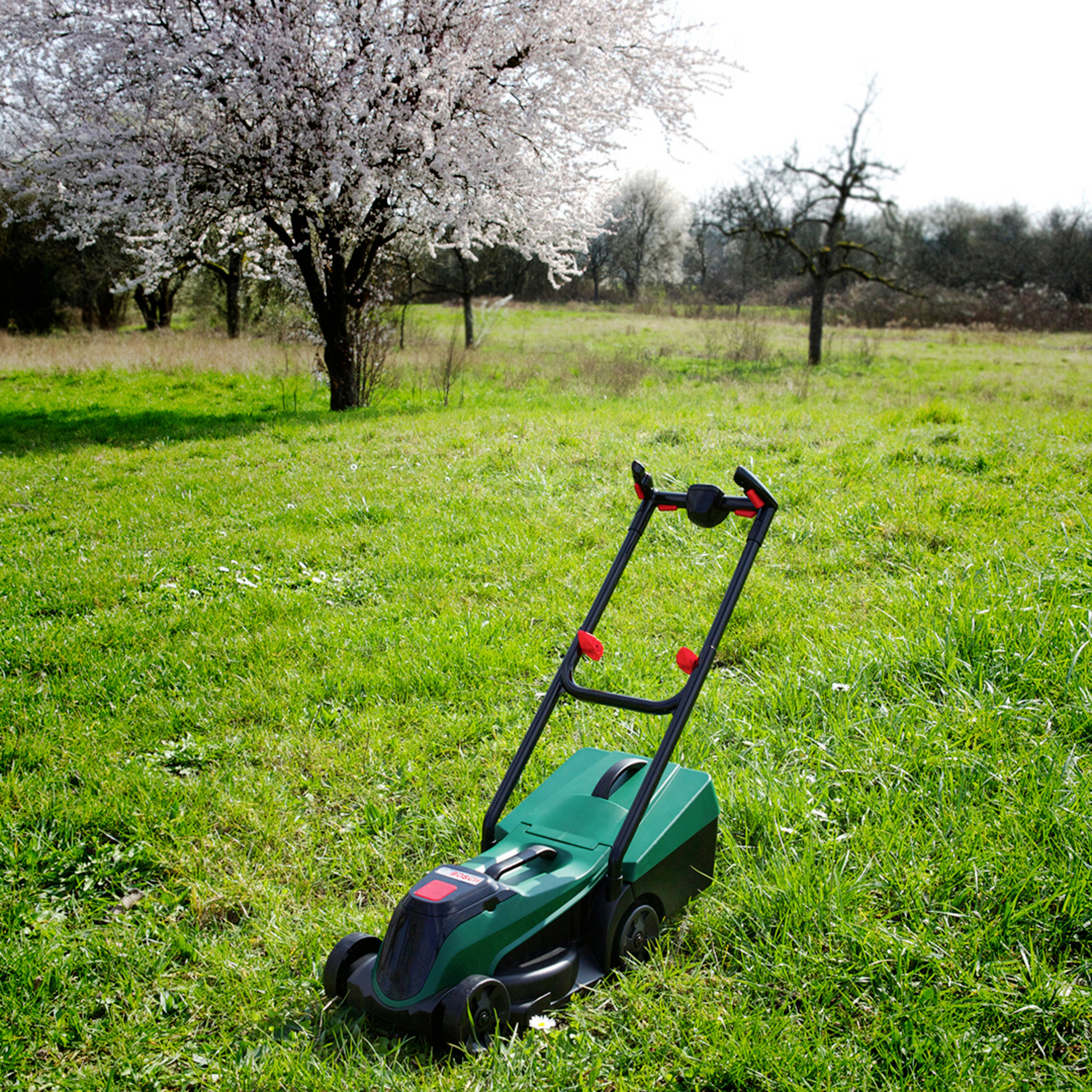A green lawn mower on vibrant grass with blooming trees in the background, suggesting a spring garden or yard maintenance scene.
