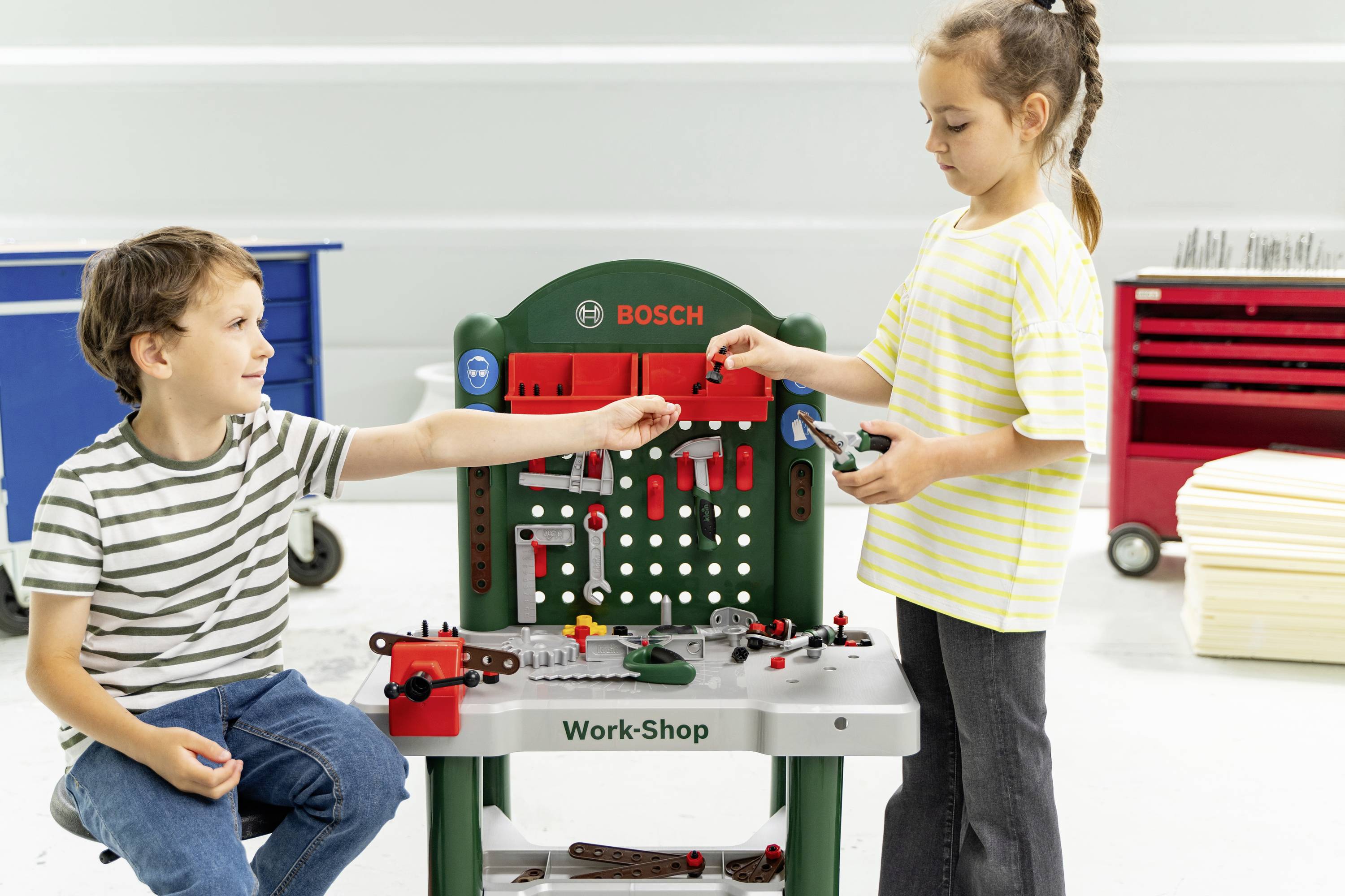 A boy and a girl are playing with a toy workbench labeled 'Work-Shop'. The boy sits holding a tool, while the girl stands, handing him a part.