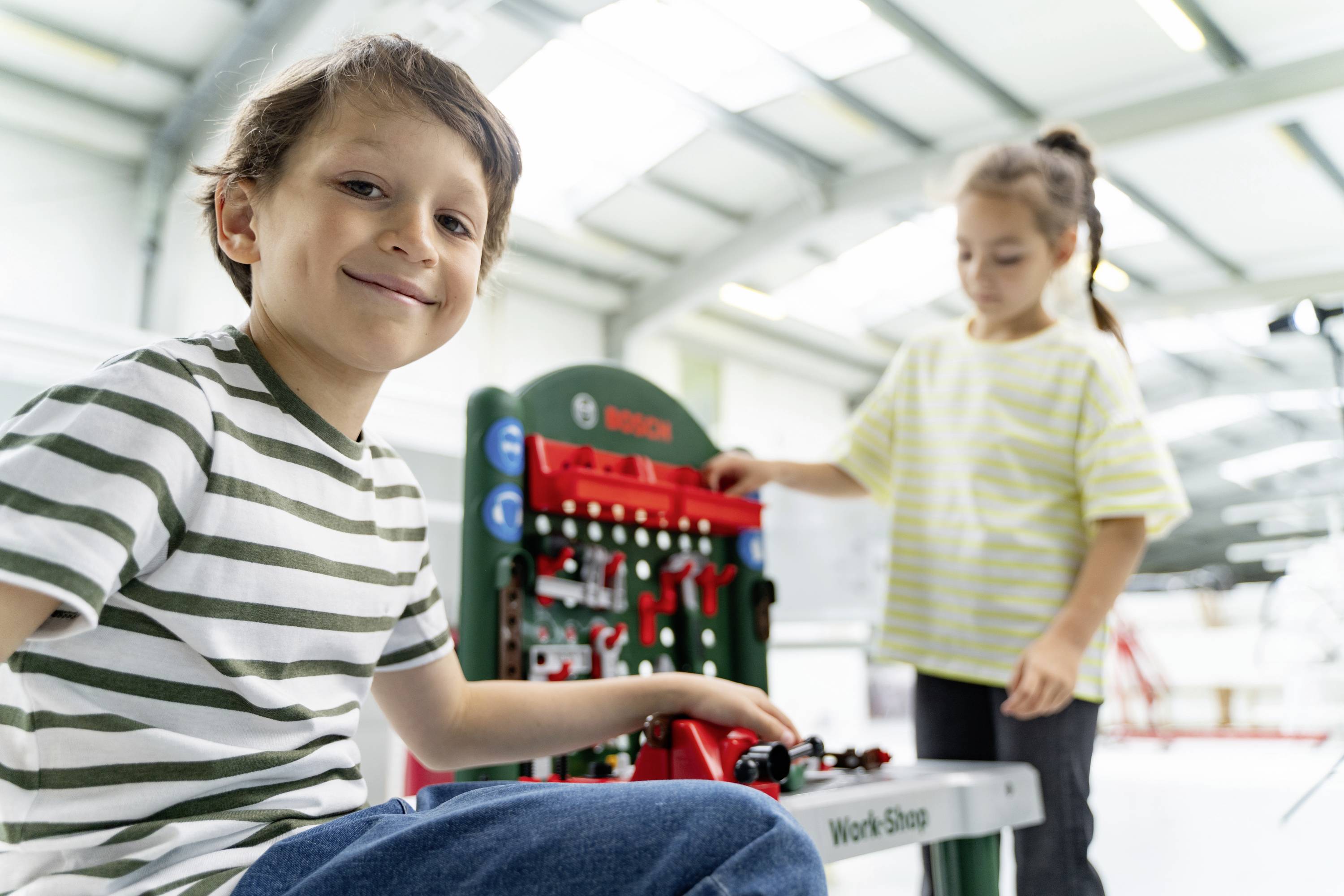 A boy and a girl play with toy tools at a workbench in a bright room. The boy smiles at the camera.