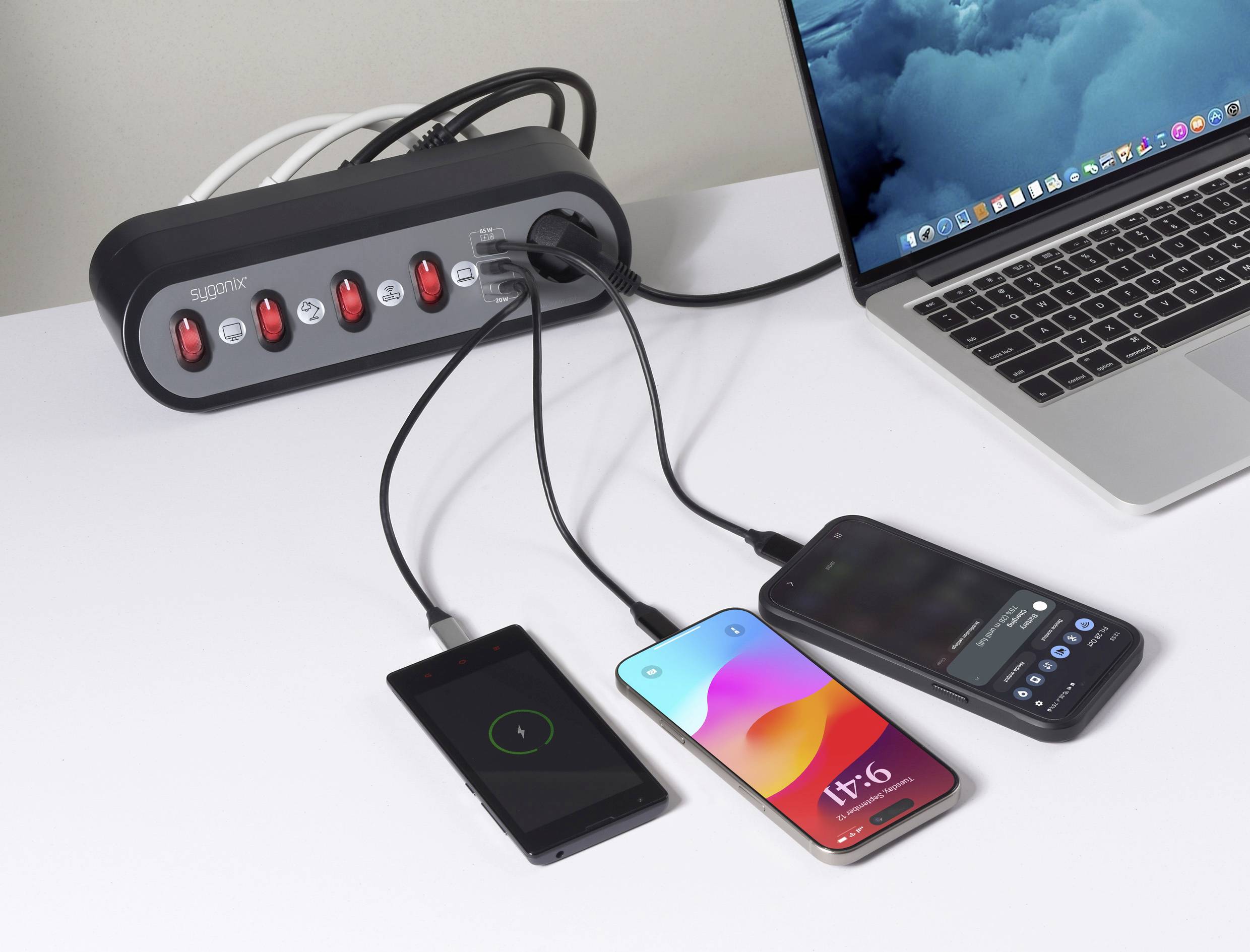 A power strip and a laptop sit on a desk. Three smartphones are connected and charging. The laptop displays a cloud-themed desktop background.