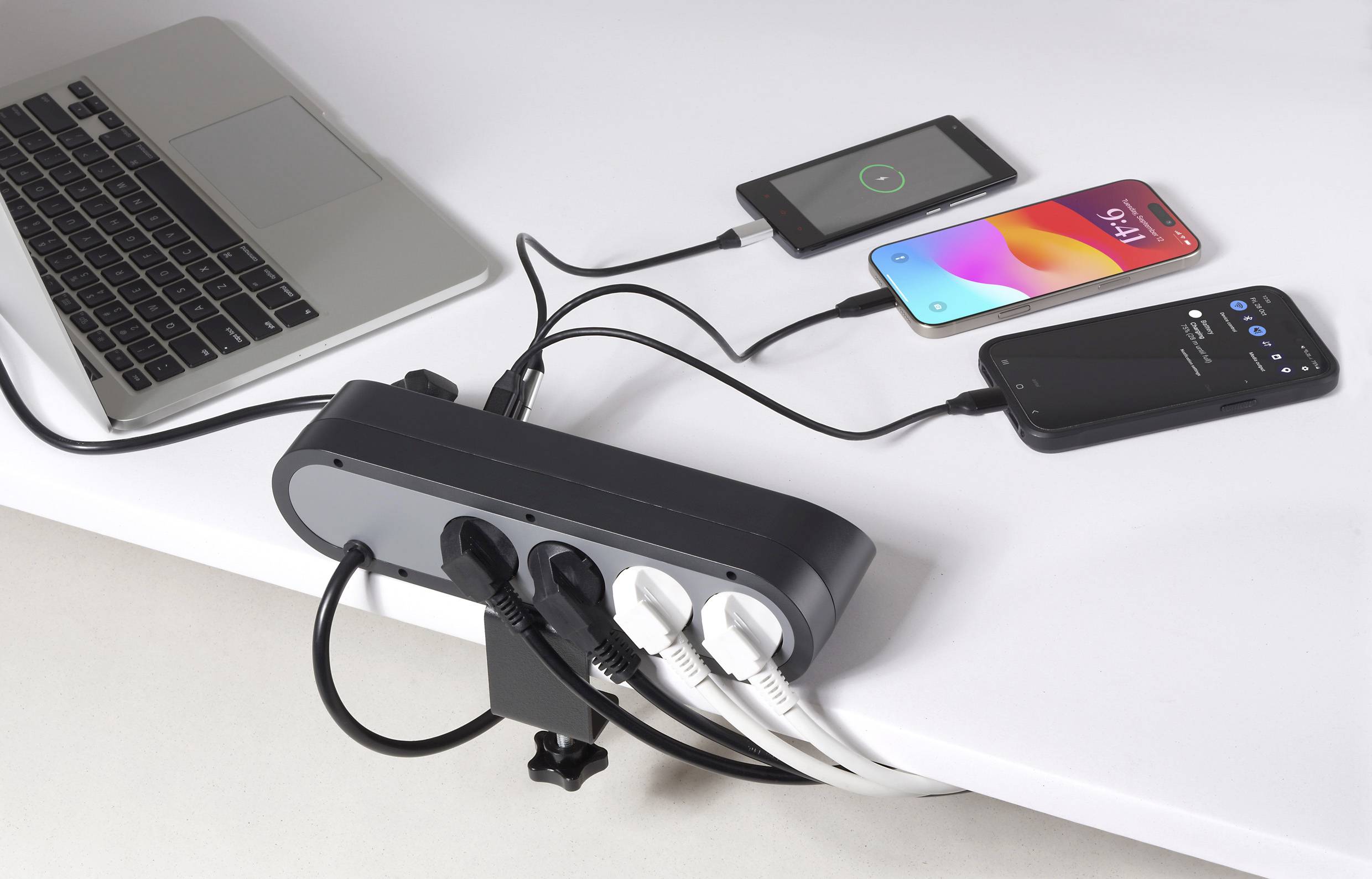 A power strip on a desk charges two smartphones and a laptop. Multiple wires connect devices, showing efficient charging setup.