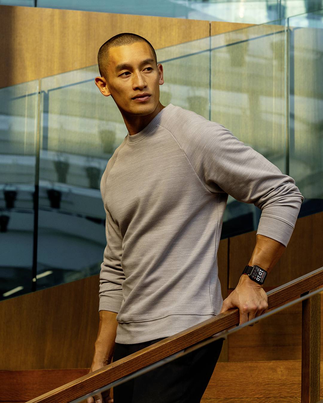 A person wearing a gray sweatshirt and a smartwatch stands on a modern staircase with a glass and wood design, looking to the side.