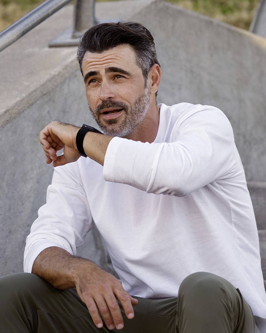 A man in casual attire, wearing a smart watch, sits on outdoor concrete steps, appearing relaxed and thoughtful.