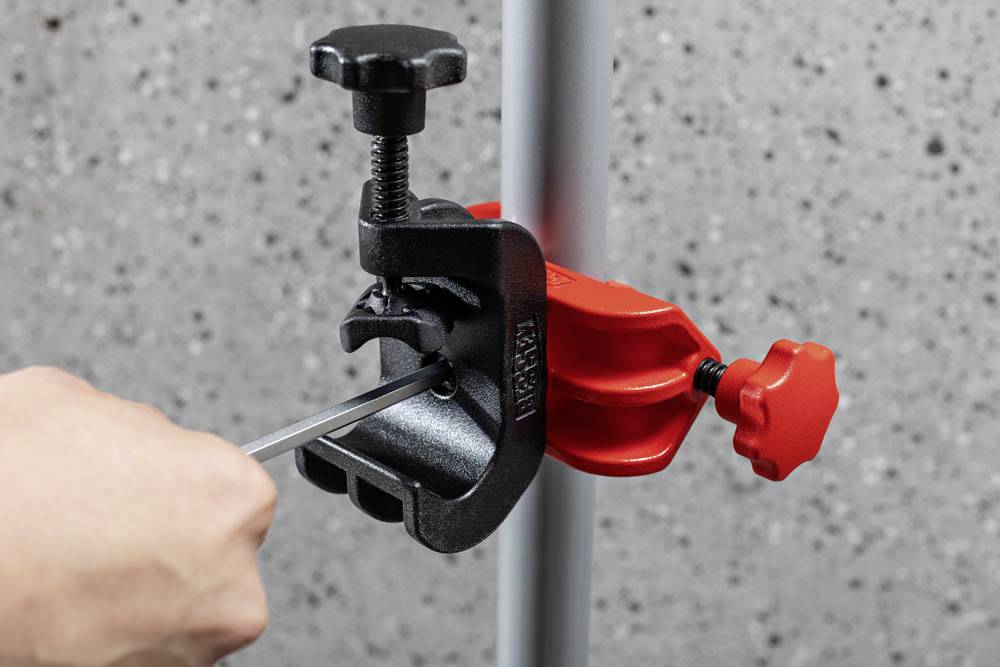 A person uses a screwdriver on a red and black metal clamp attached to a vertical pole against a concrete wall background.