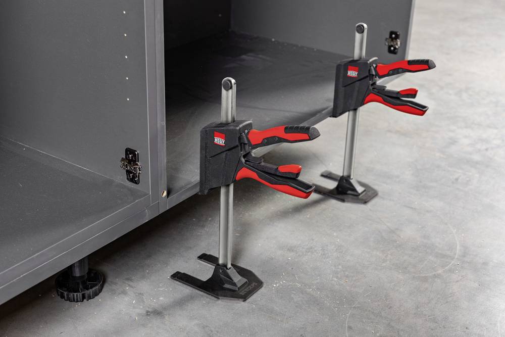 Two red and black clamps are securing a metal bar between two grey cabinets on a concrete floor.