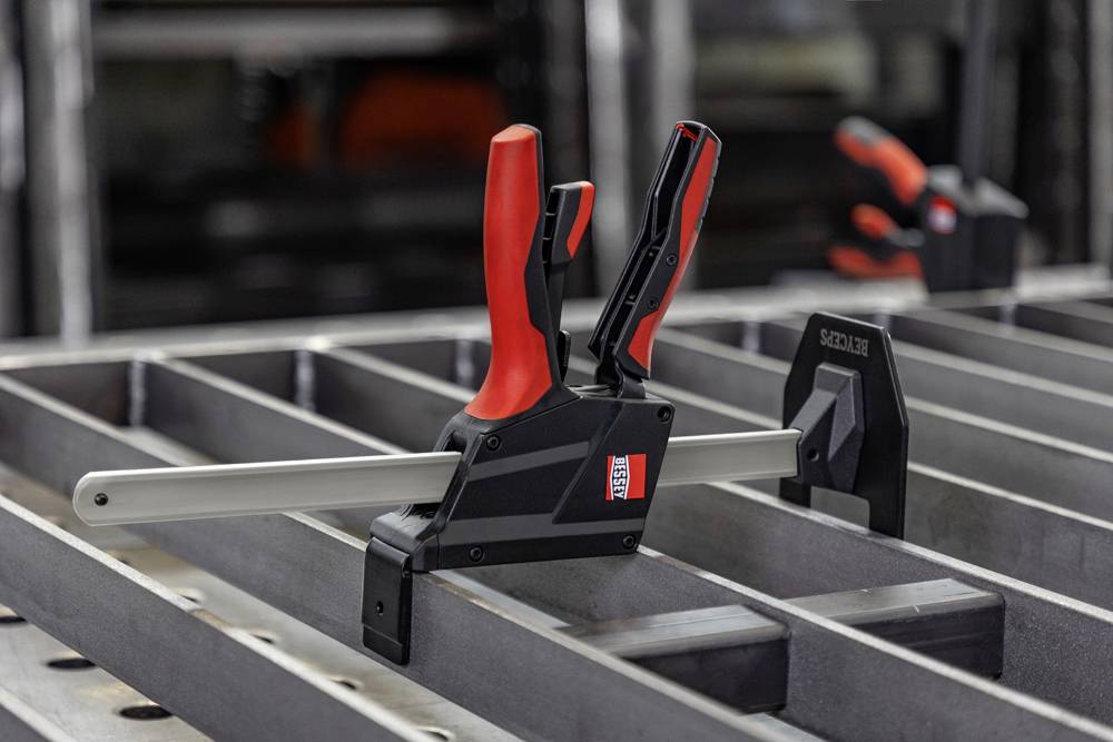A metal clamp with red handles secures a steel beam on an industrial workbench, with another clamp in the background.