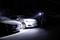 A security guard inspects a row of parked cars in a dimly lit parking lot, using a flashlight for close examination.