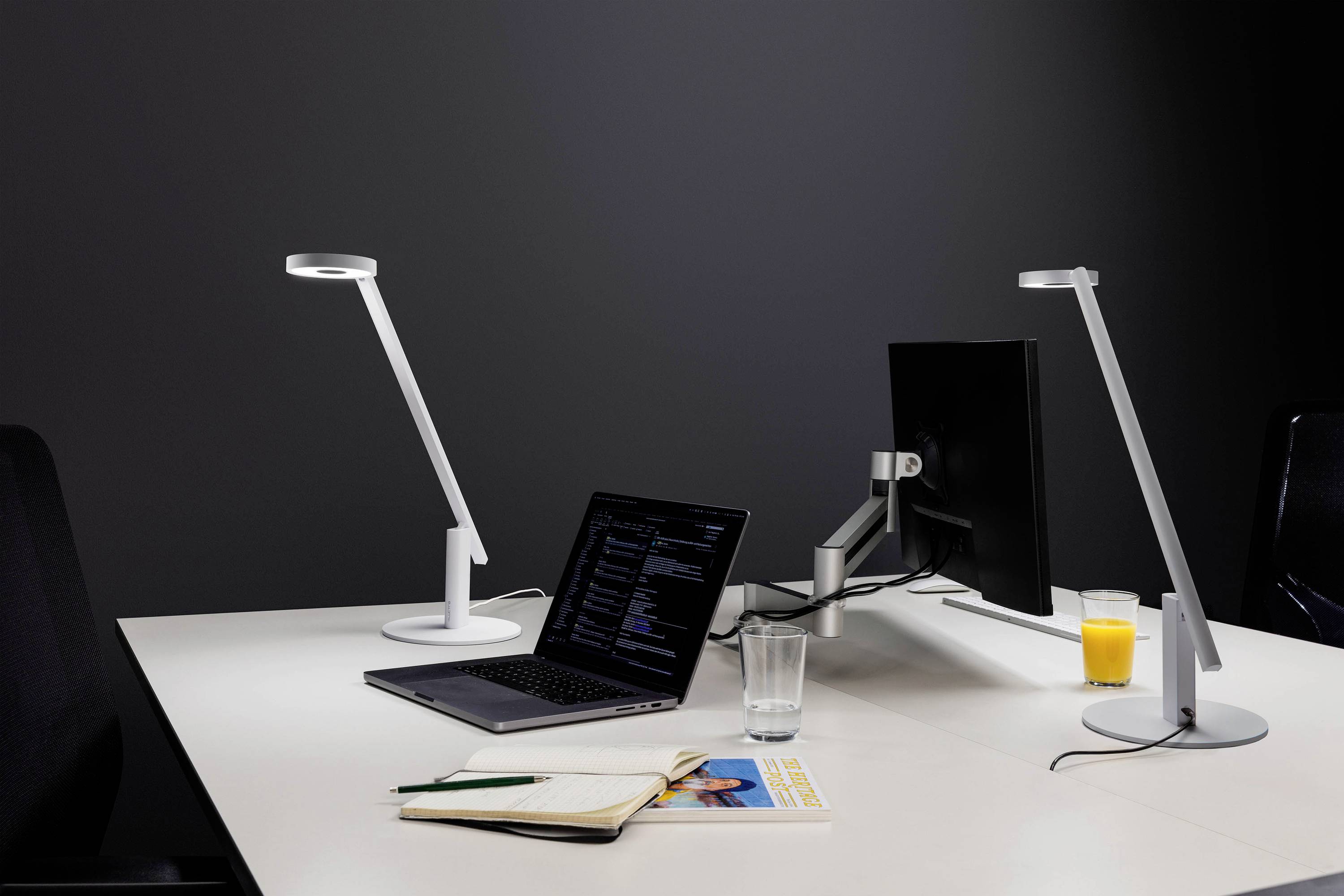 A modern workspace featuring a laptop displaying code, a monitor, desk lamps, an open notebook, a glass of water, and a glass of orange juice.
