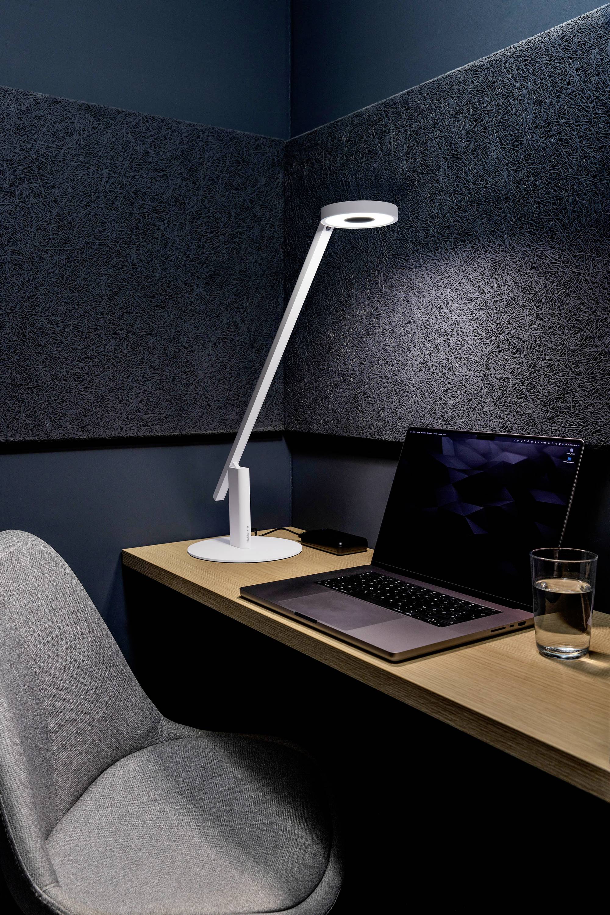 A minimalist workspace with a grey chair, wooden desk, open laptop, desk lamp, and glass of water in a dark, padded room.