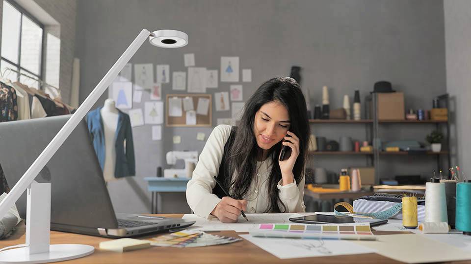 A woman in a design studio talks on the phone while sketching at a desk. Fabrics, a laptop, and design materials are visible.