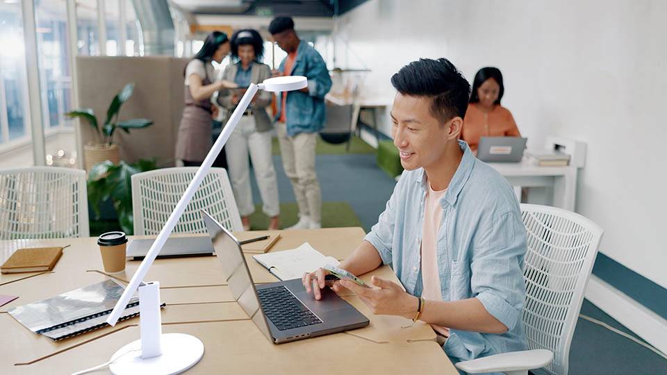 A person working at a laptop in a bright, modern office, holding a smartphone, while three colleagues discuss papers in the background.