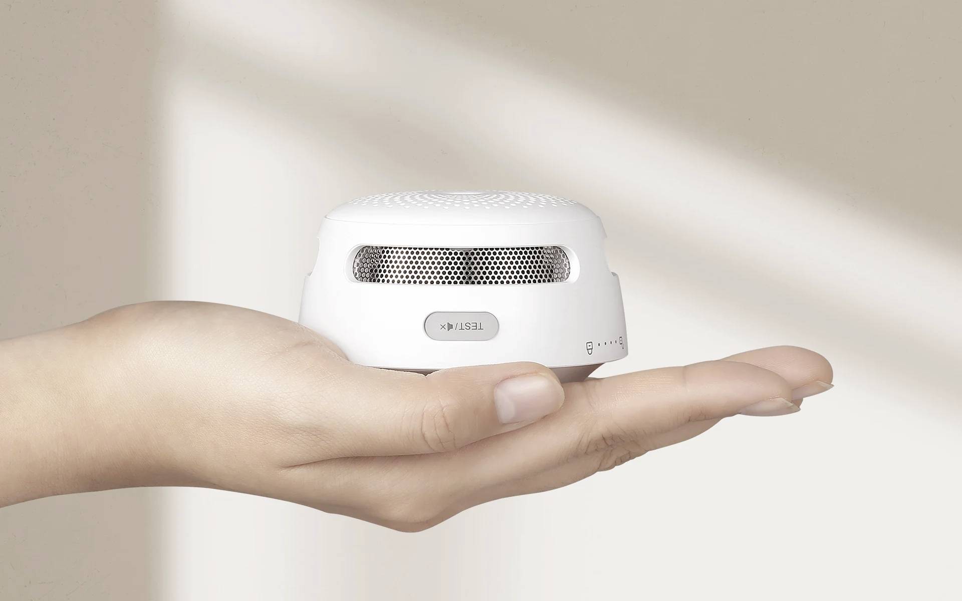 A hand is holding a small, round, white smoke detector. The background is blurry and beige.