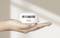 A hand is holding a small, round, white smoke detector. The background is blurry and beige.