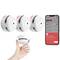 A hand holds a white smoke detector. Nearby, three identical detectors, and a smartphone showing an app alert 'Smoke Alarm Triggered'.