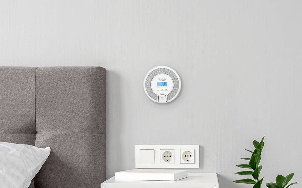 A smart thermostat displaying temperature and settings is mounted on a gray wall above a headboard and beside a white power outlet.