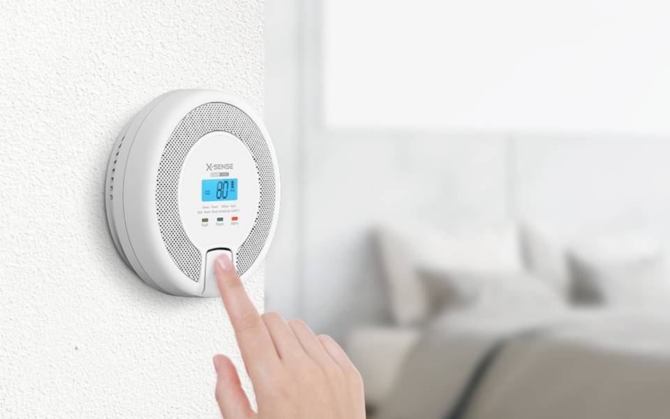 A hand pressing a button on a wall-mounted smoke detector with a digital display showing '80.1'. A blurred bedroom is in the background.