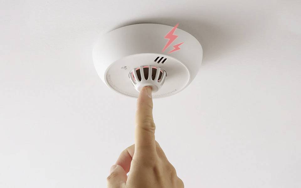 A person presses a button on a ceiling-mounted smoke detector, depicted with red lightning symbols indicating activation or testing.