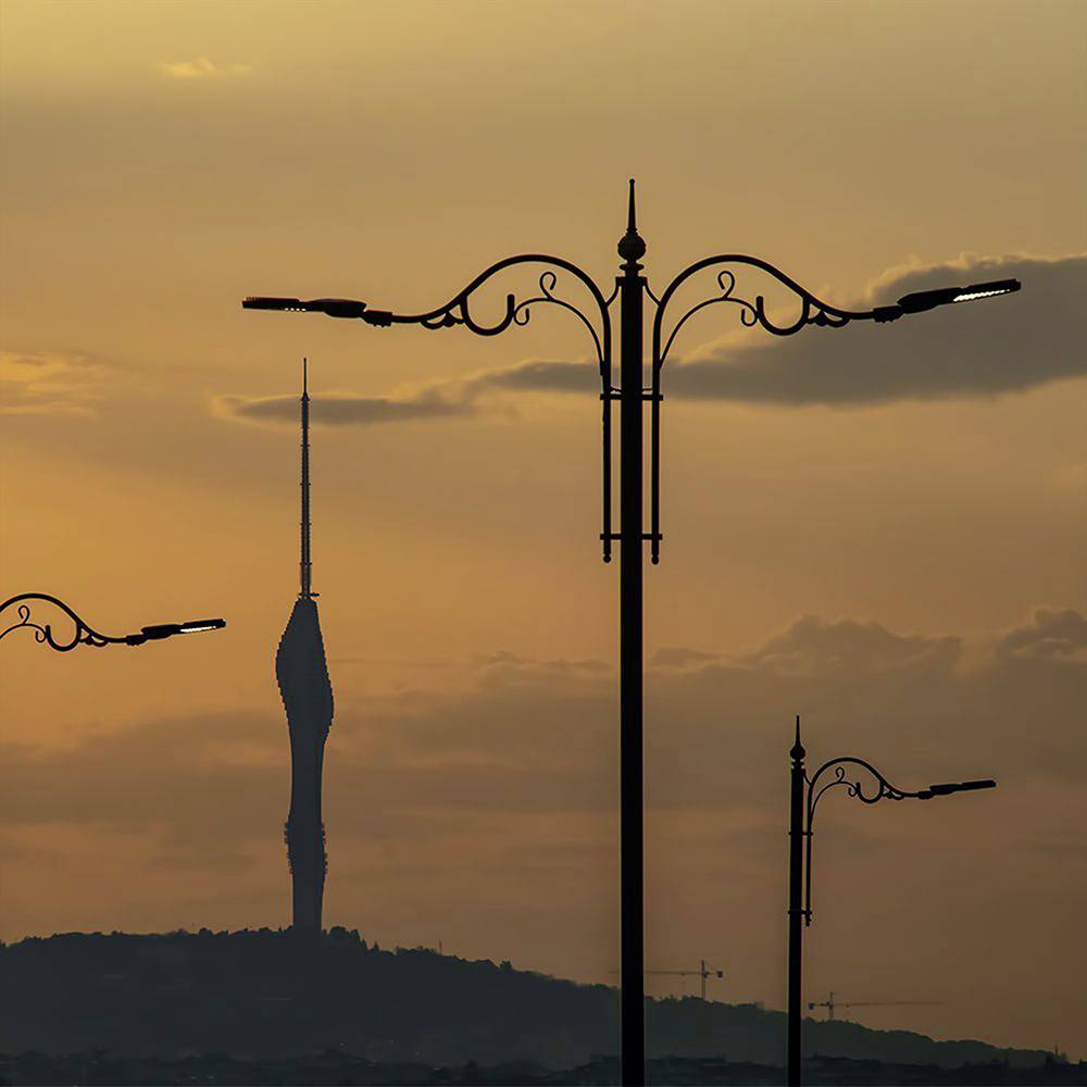 A tall, slender tower stands silhouetted against an orange sunset sky, with ornate streetlights in the foreground creating a detailed skyline.