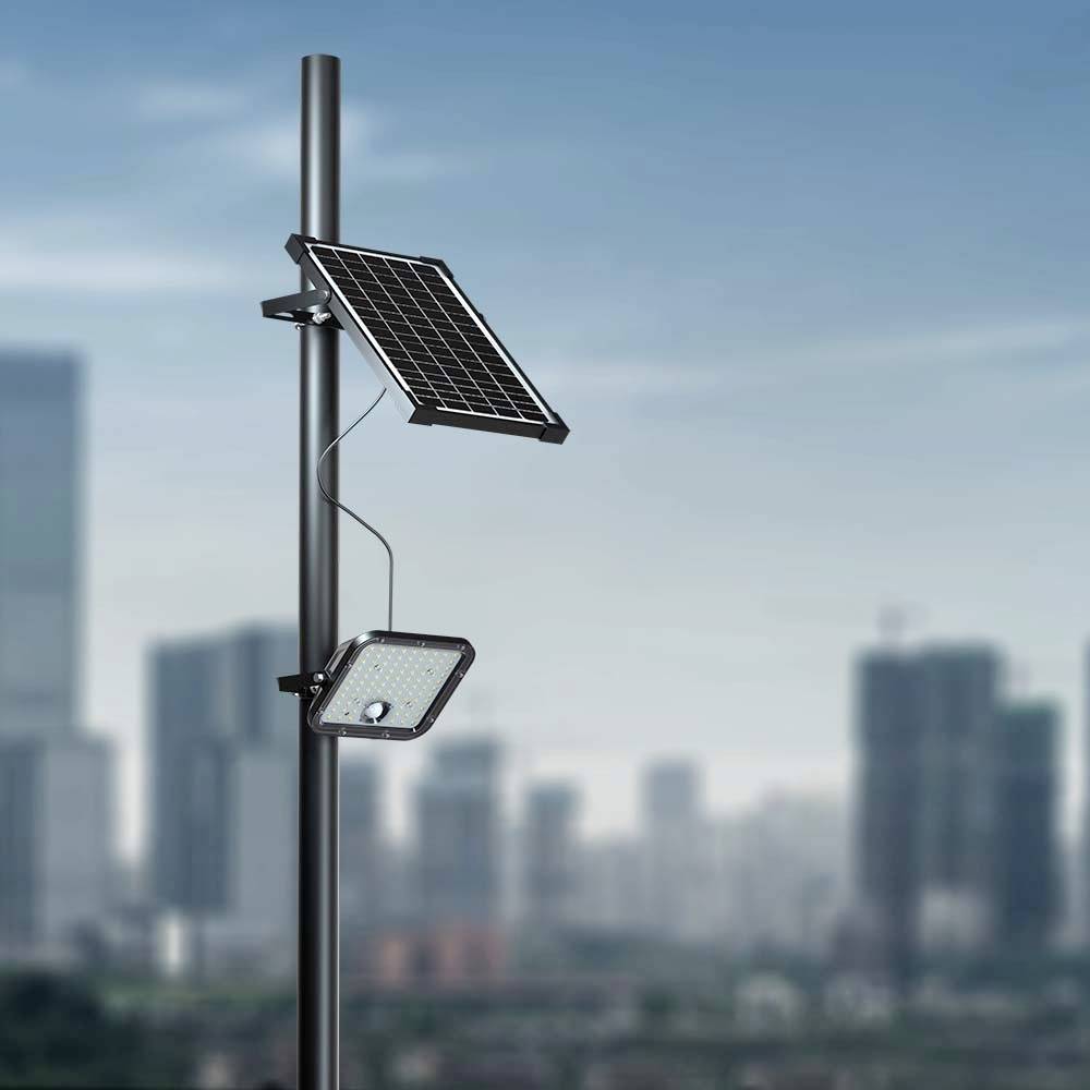 A streetlight with a solar panel stands tall against a cityscape backdrop of skyscrapers under a clear blue sky.
