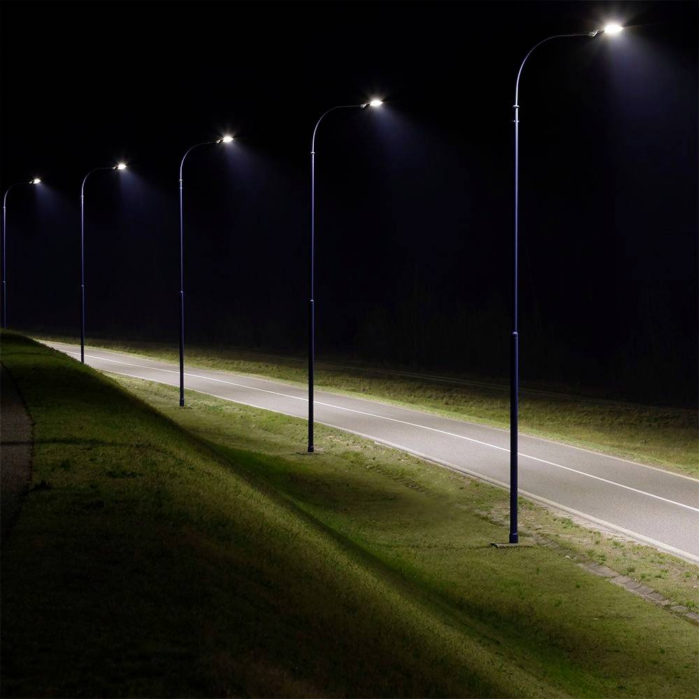 A row of streetlights illuminates an empty asphalt road at night, casting light on grassy areas on either side.