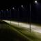 A row of streetlights illuminates an empty asphalt road at night, casting light on grassy areas on either side.