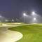 A night golf course illuminated by bright lights with lush green fairways and sand bunkers, surrounded by palm trees.