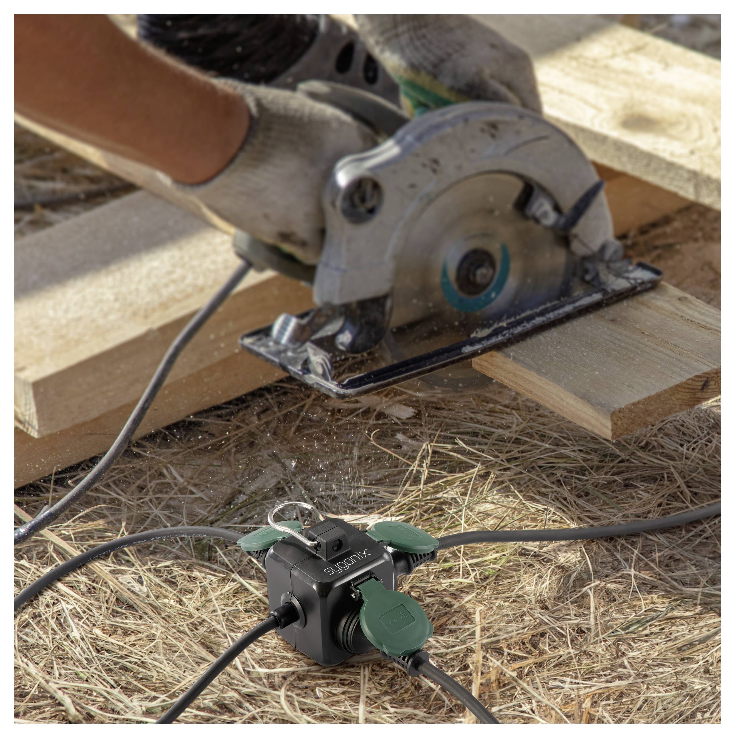 A person wearing gloves uses a circular saw to cut wooden planks on the ground. An electrical power strip with cords is visible nearby.