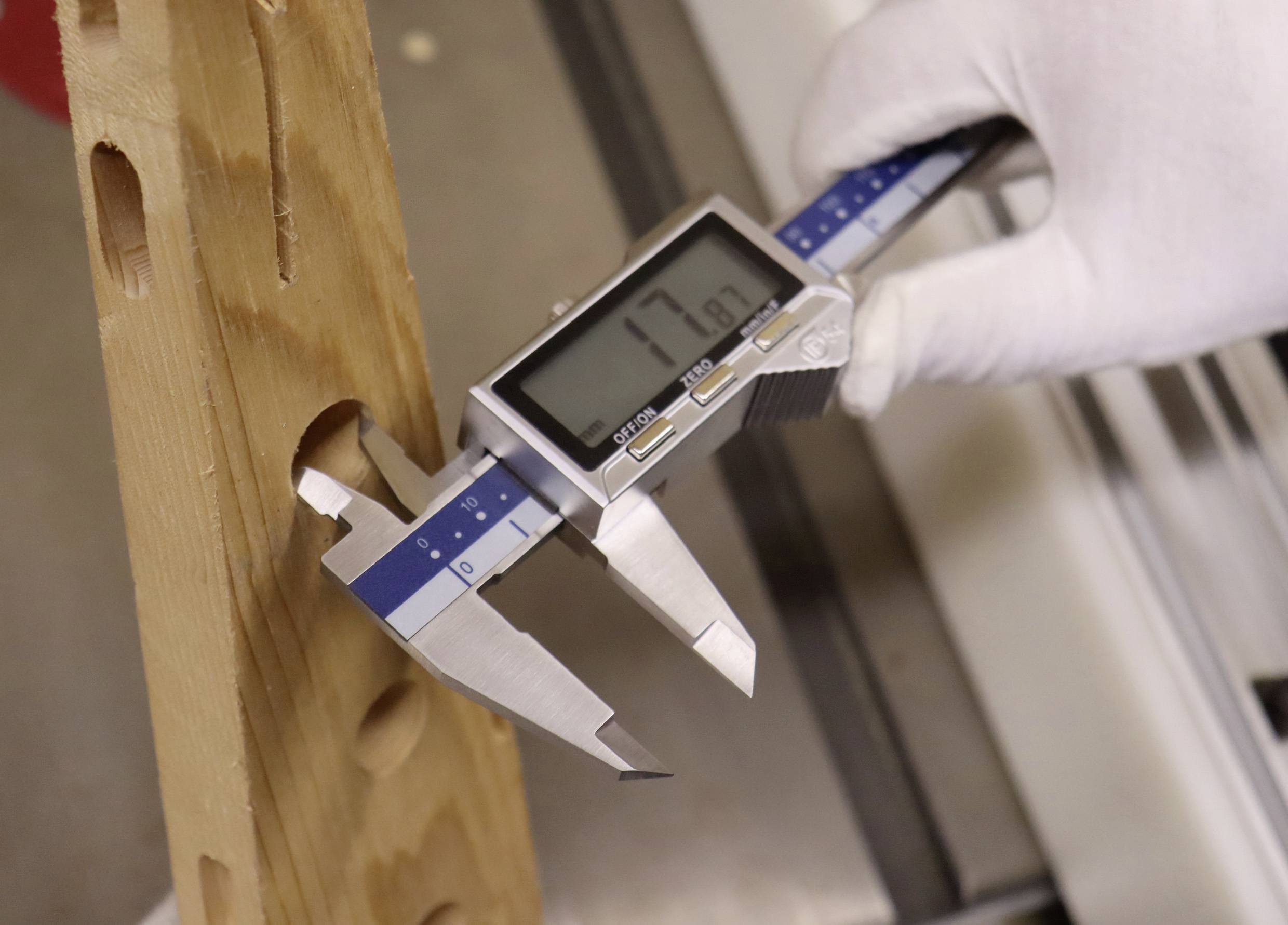 A digital caliper measuring the thickness of a wooden block, displaying '17.1 mm' on the screen, held by a gloved hand.