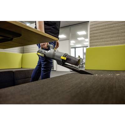 A person uses a handheld vacuum to clean crumbs from a dark-colored couch in a modern office setting.