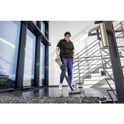 A person is vacuuming a modern building's staircase landing with a cordless vacuum cleaner. The area features large windows and metal railings.