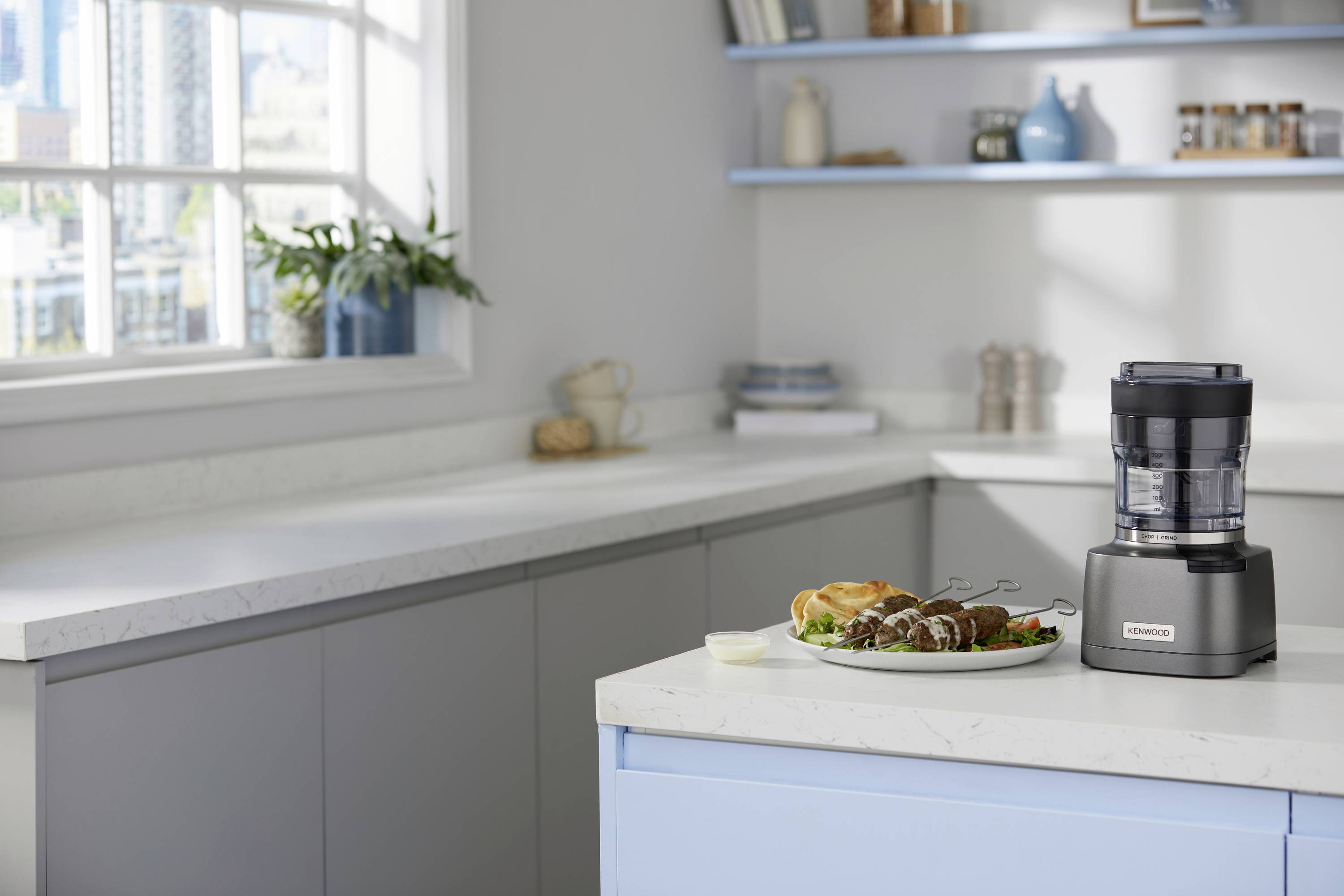 A kitchen with a food processor on the countertop next to a plate of skewered meat and vegetables, with bright natural light.
