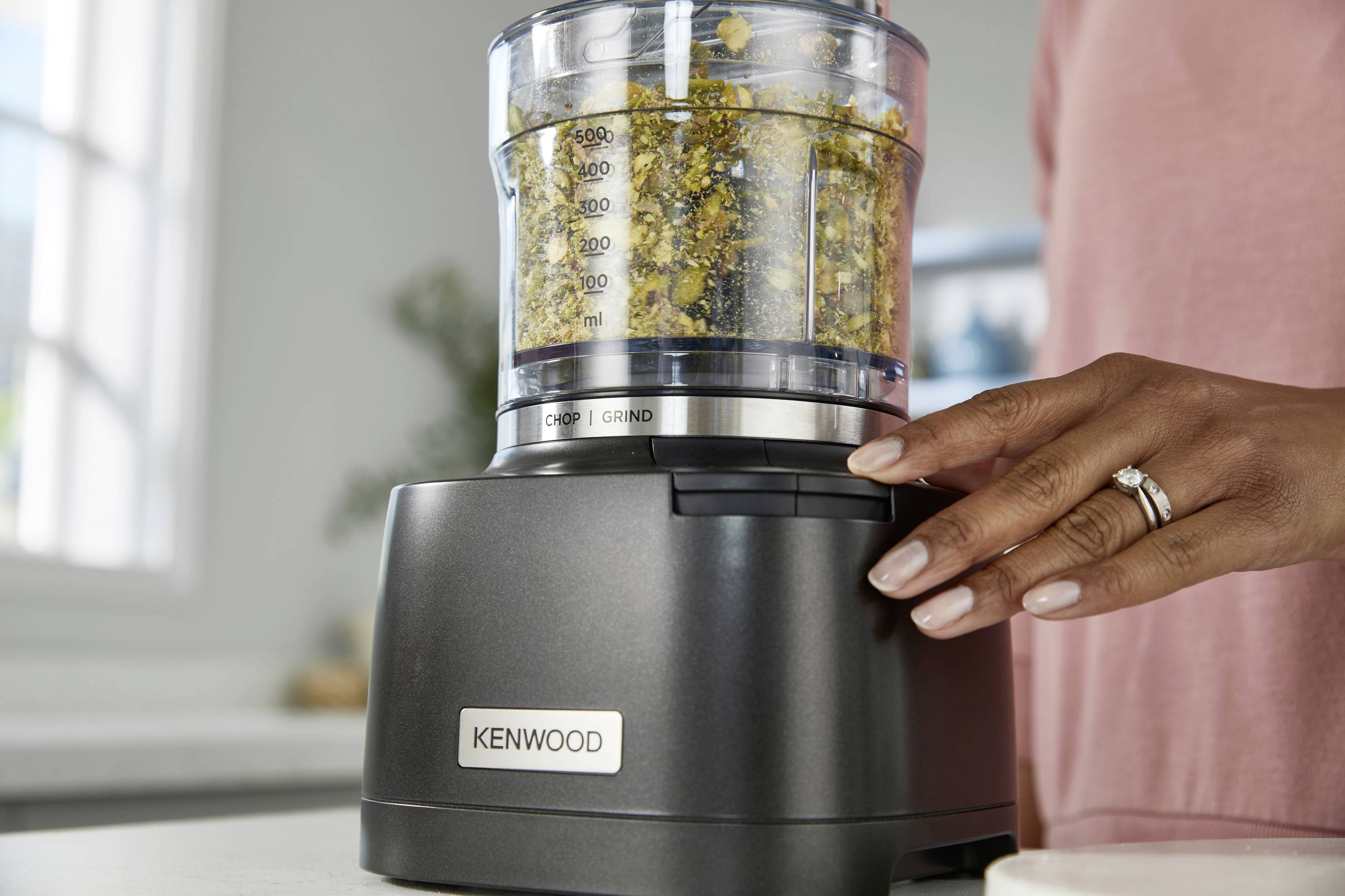 A hand operates a Kenwood food processor filled with herbs, in a bright kitchen setting.