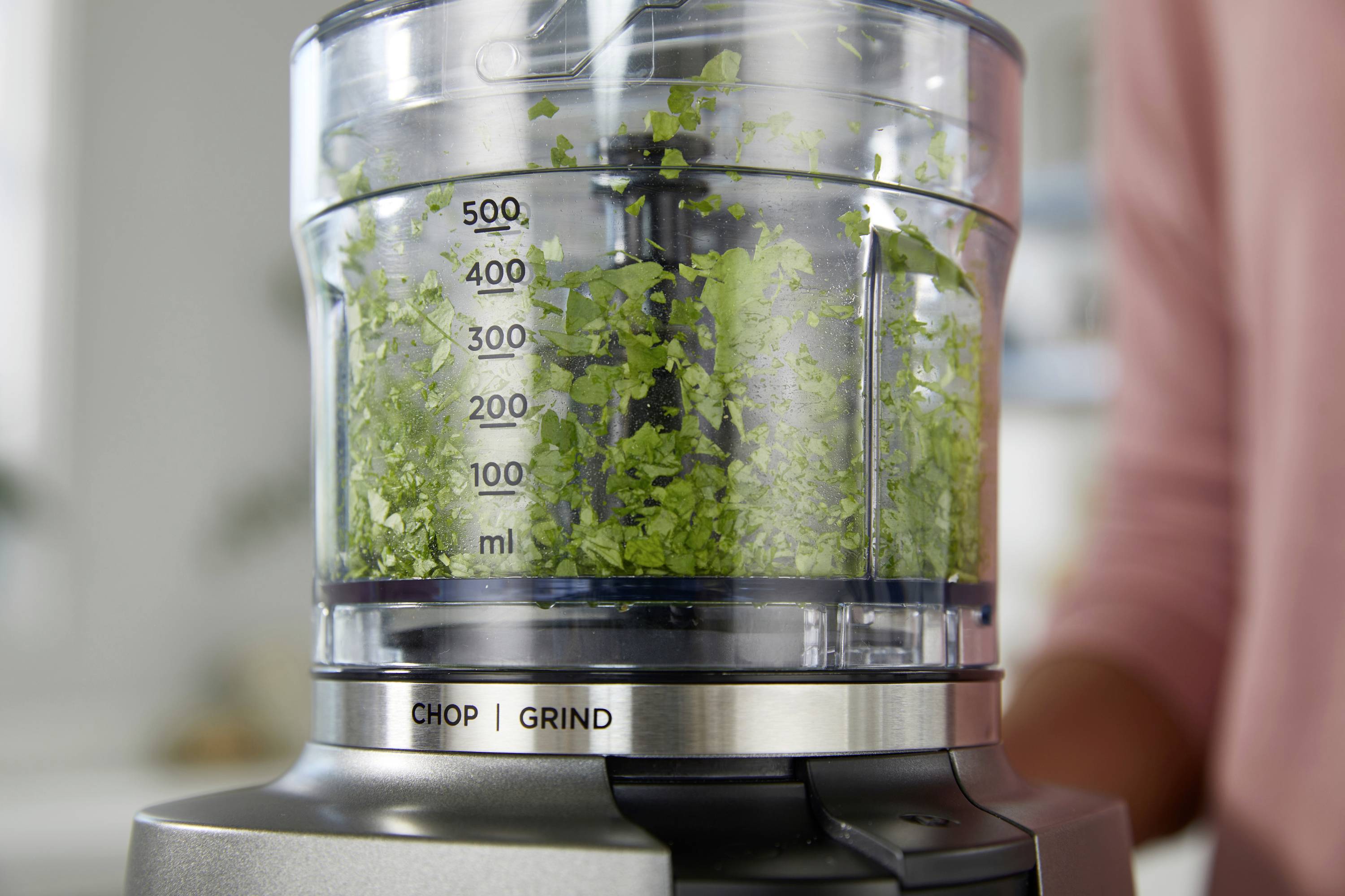 A close-up of a blender processing green herbs, with measurements on the container. The blender has settings labeled 'CHOP' and 'GRIND'.