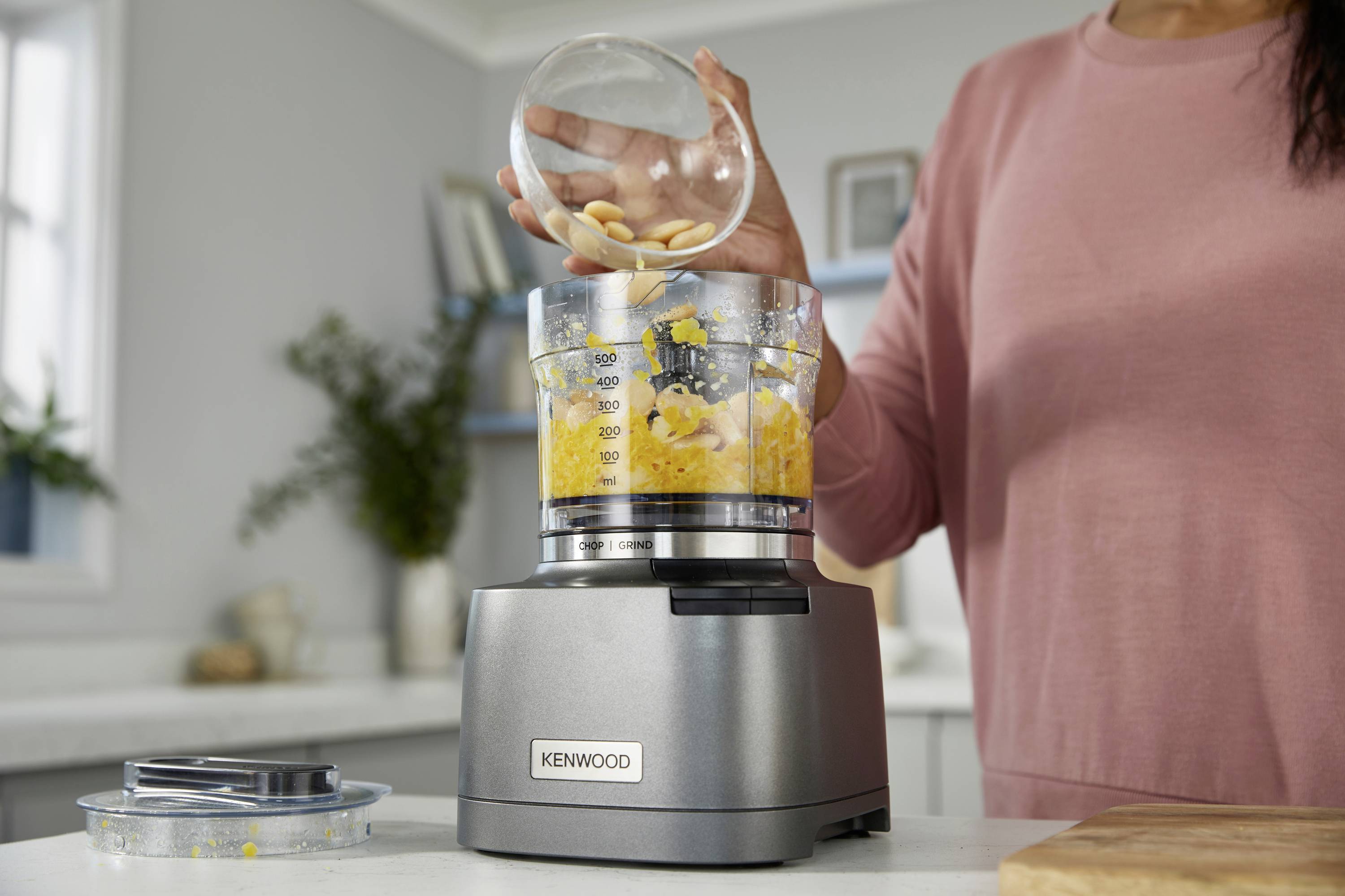 A person adds ingredients to a food processor blending a mixture, in a modern kitchen setting.
