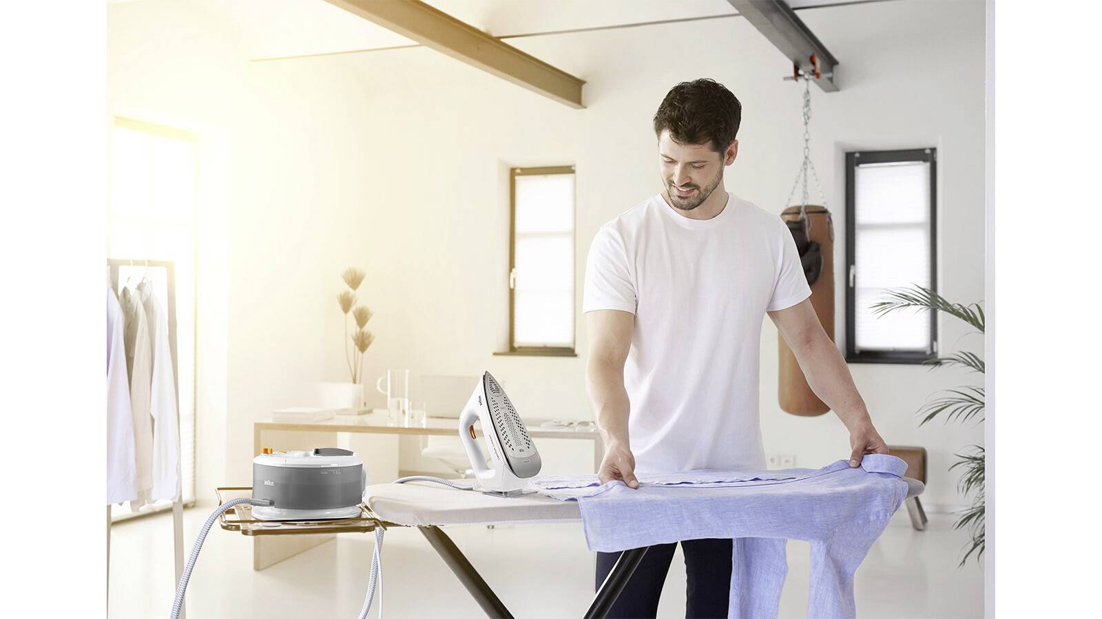 A man in a white shirt irons a blue shirt on an ironing board in a bright, modern room with windows and a punching bag in the corner.